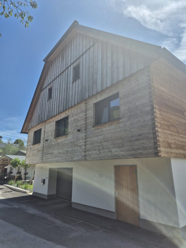 Fassade aus naturbelassener Lärche mit gehackter und gebürsteter Blockwandschalung, traditioneller Klingschrotverbindung und sägerauer Lärchenschalung im steirischen Stil.